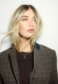 Ruda ir žalia houndstooth tipo striukė su mėlynu apykakle, apsivilkta tamsiu marškinėliu ir derinama su auksiniu grandininiu kaklo papuošalu, turinčiu pakabuką.