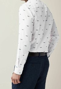 Camisa blanca de manga larga con un pequeño patrón de libélulas azules, que presenta un cuello con botones y puños en contraste, combinada con pantalones oscuros.