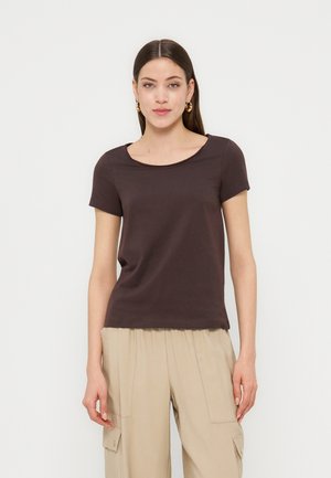 Femme portant un haut à manches courtes marron foncé et un pantalon cargo beige, debout devant un fond clair uni.