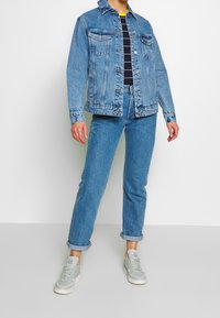 Denimová bunda a odpovídající džíny v světle modré barvě. Bunda má náprsní kapsy s klopami, zapínání na knoflíky a vyhrnuté rukávy. Nošeno s šedými teniskami.