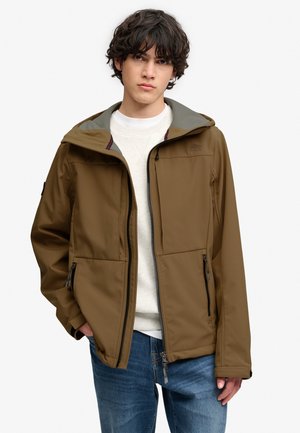 Junger Mann mit lockigem dunklem Haar, der eine braune Jacke mit Reißverschluss über einem weißen Pullover und blauen Jeans trägt, steht mit einer Hand in der Tasche.