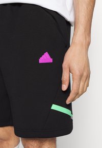 Shorts noirs avec un logo Adidas rose et une bande horizontale verte près de l'ourlet, portés par une personne avec une main visible et une chemise blanche.