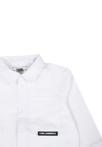 Camicia bianca a maniche corte con chiusura a bottoni, colletto a punta, taschino sul lato sinistro e un'etichetta nera con la scritta "KARL LAGERFELD" in testo bianco.