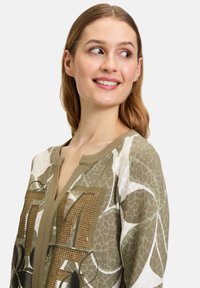 Grön och vit mönstrad blus med rund halsringning, metallstudsar på framsidan och en avslappnad passform med långa ärmar.