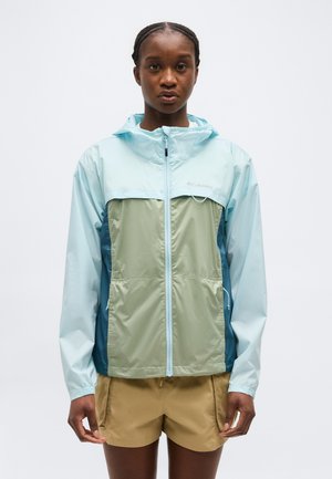 Naine kannab helerohelist ja rohelist tõmblukuga kapuutsiga jopet ning beeže lühikesi pükse, seisab ühevärvilise heleda tausta ees.