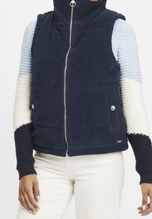 Person iført en navyblå polstret vest med lynlås over en hvid og navy ribstrikket sweater og hvide bukser, stående mod en ensfarvet baggrund.