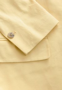 Žlutá bavlněná látka blejzru s klopou a detailem kapsy. Obsahuje kulatý šedý knoflík se čtyřmi otvory.