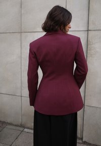 Bordinės pritaikytos švarkas su struktūruotais petimis, aštriu apykakle ir vertikaliomis siūlėmis; derinamas su ilgomis juodomis kelnėmis priešais betono foną.