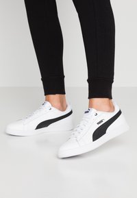 Weiße Sneakers mit schwarzem Akzentstreifen, glattem Synthetik-Obermaterial, runder Zehenpartie, Schnürdesign und strukturiertem Gummisohle. Puma-Logo an der Seite.