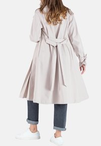 Lichtbeige trenchcoat met een tailleriem, met een uitlopende snit en zijinsneden. Gepaard met opgerolde donkere jeans en witte sneakers.