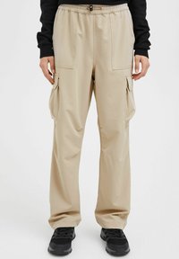 Pantalons cargo beiges avec une taille élastique, dotés de deux poches latérales et d'un design de jambe fuselée. Portés avec des baskets noires.