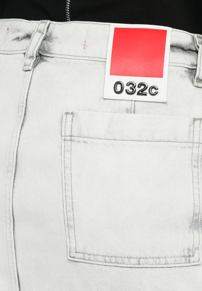 Pantalon en denim gris clair avec une étiquette rouge portant le texte "032c". Détails notables comprenant une poche arrière et des coutures visibles.
