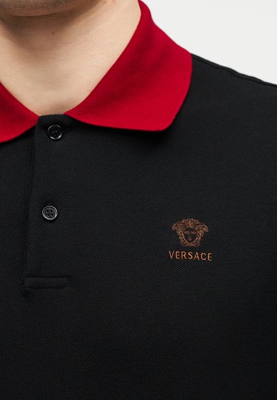 Εγγύς πλάνο ενός μαύρου πόλο πουκαμίσου με κόκκινο γιακά, μαύρα κουμπιά και μικρό κεντημένο λογότυπο Versace στο στήθος.