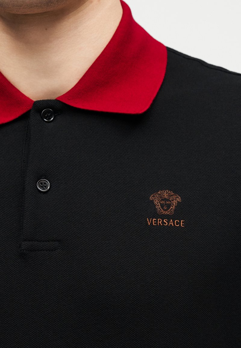 Close-up of a black polo shirt with a red collar, black buttons, and a small embroidered Versace logo on the chest.