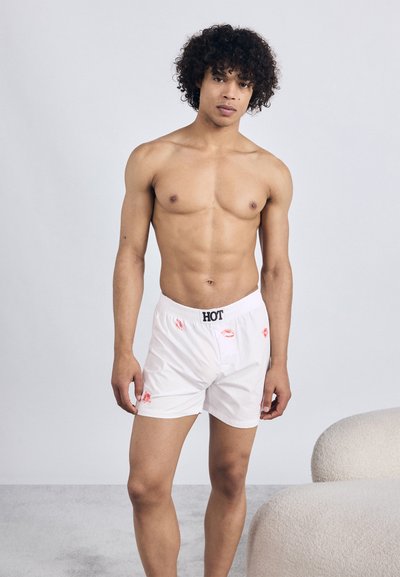 Boxeri albi cu imprimeu de buze roșii, talie neagră cu „HOT”, din material de bumbac, croială lejeră și fundal simplu. Modelul are părul creț.