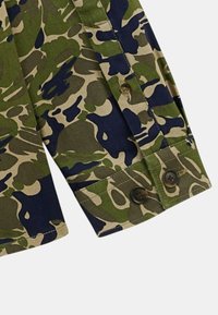 Manche de chemise camouflage avec des motifs verts, bleu marine et beige. Comprend des boutons marron et un poignet replié pour un détail supplémentaire.