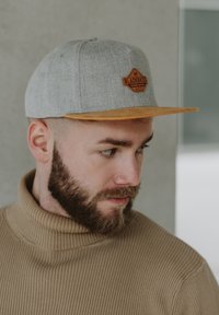 Grå tygsnapbackkeps med en platt orange skärm och en brun läderlapp märkt "BLACKSKIES." Modell bär en rippad beige polotröja.