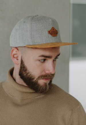 Siva tkanina snapback kapa s ravnim narančastim vizirima i smešnim kožnim oznakom na kojoj piše "BLACKSKIES." Model nosi rebrasti bež rolak.