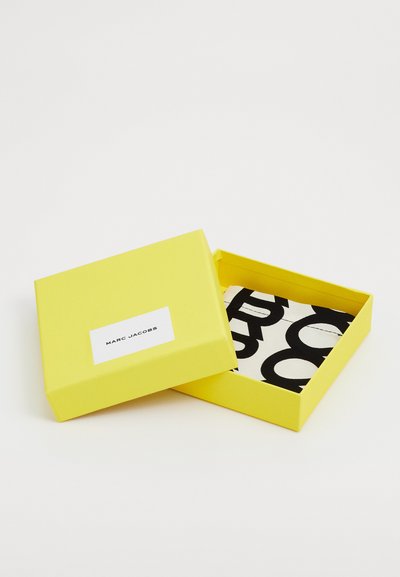 Gul papkasse med en hvid etiket, der viser "MARC JACOBS." Indeni er der en sort-hvid mønstret stof. Æsken er åben.