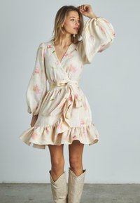 Blumenmuster-Wickelkleid in hellem Beige mit rosa Akzenten. Mit langen Puffärmeln, einem Taillengürtel und einem gerafften Saum. Cowboy-Stiefel werden getragen.