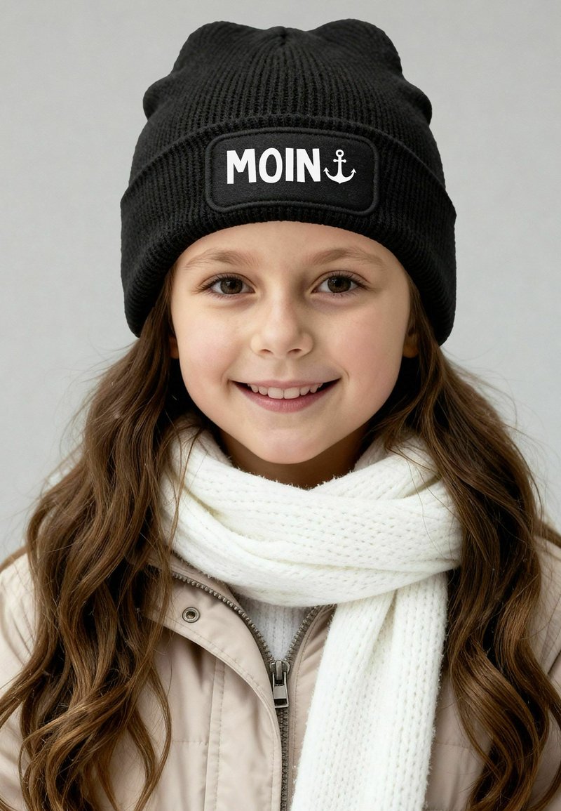 Black knitted beanie with a white rectangular patch displaying "MOIN" and an anchor symbol. Worn with a white textured scarf and light jacket.