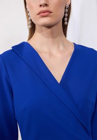Sininen kietaisumallinen mekko, jossa on V-pääntie; valmistettu sileästä kankaasta. Siinä on kevyt kaulus ja se on yhdistetty roikkuviin hopeakorvakoruisiin.