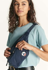 Bolso bandolera de nailon azul marino con textura acolchada, cierre de cremallera y parche redondo con logo. Correa ajustable llevada sobre una camisa azul clara.