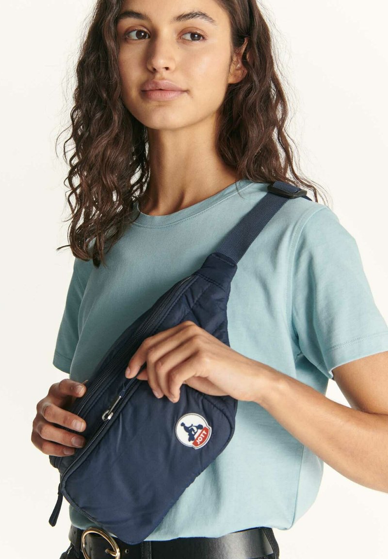 Bolso bandolera de nailon azul marino con textura acolchada, cierre de cremallera y parche redondo con logo. Correa ajustable llevada sobre una camisa azul clara.