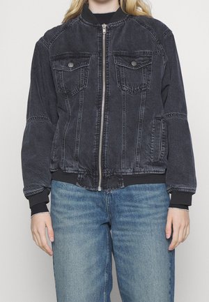 Giacca bomber in denim nero con colletto e polsini a coste, dotata di due tasche sul petto e chiusura con zip. Indossata con jeans azzurri chiari.