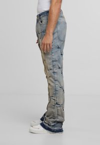 Jasnoniebieskie dżinsy o tapered fit, z marszczoną teksturą i postrzępionymi brzegami, zestawione z białymi sneakersami.