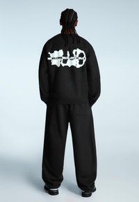 Čierna mikina s veľkým bielym textom "WORLDWIDE PLAYGROUND" na zadnej strane, skombinovaná s voľnými čiernymi nohavicami. Hladká textúra látky.