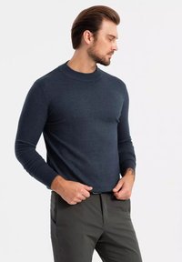 Dunkelblauer Rundhalsausschnitt-Pullover mit langen Ärmeln, aus glattem Strickstoff, kombiniert mit dunkelgrünen Hosen, vor einem schlichten Hintergrund stehend.