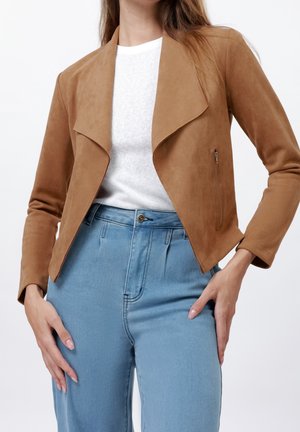 Kvinde iført en lysebrun ruskindskort jakke over en hvid top med lyseblå højttaljede jeans, hænderne hvilende på hofterne.