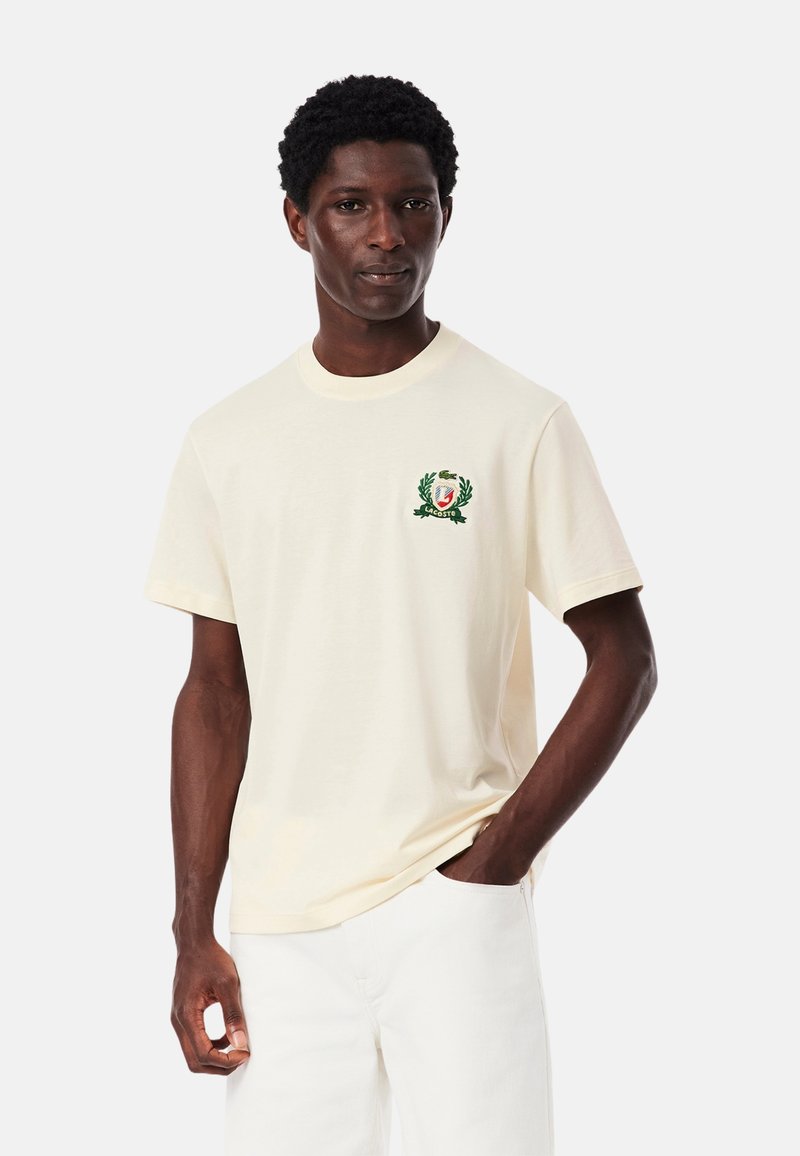 T-shirt di cotone color crema con logo ricamato verde e design di corona d'alloro. Maniche corte e scollatura rotonda.