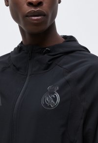 Κοντινό πλάνο ενός ατόμου που φοράει μαύρο μπουφάν με κουκούλα, με μονοχρωματικό λογότυπο της Ρεάλ Μαδρίτης στο στήθος, πάνω σε απλό φόντο.