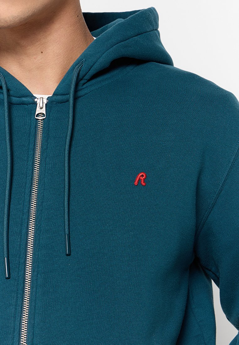 Türkise Zip-Hoodie mit rotem gesticktem Logo. Verfügt über eine Kapuze, einen vorderen Reißverschluss und eine weiche Stofftextur. Klassische Passform.