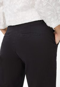 Schwarze Hosen mit glatter Textur, ausgestattet mit zwei Gesäßtaschen und einem schlanken Schnitt. Kombiniert mit einer weißen, bestickten Bluse.