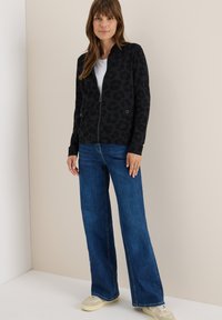 Veste noire zippée avec imprimé léopard, poignets côtelés et poches latérales, associée à un jean large bleu et des baskets beiges.