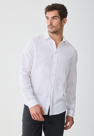 White linen long-sleeve shirt with a button-down collar, featuring a relaxed fit and slightly textured fabric, paired with dark trousers.
