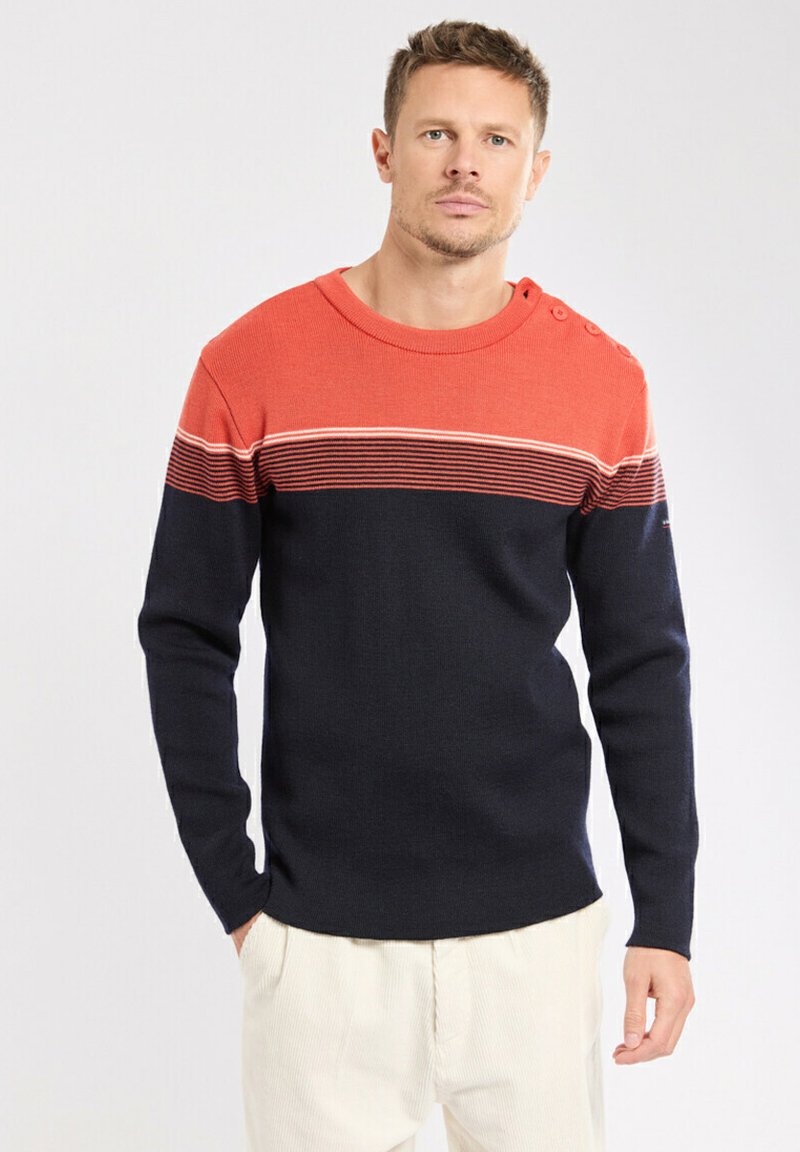 Homme portant un pull à manches longues avec des rayures horizontales orange et noires, se tenant les mains dans les poches, sur un fond blanc.
