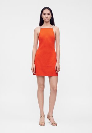 Femme aux longs cheveux noirs portant une mini-robe sans manches orange vif et des sandales à talons hauts dorées, debout devant un fond blanc uni.