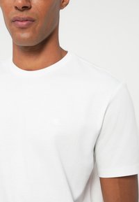 Nahaufnahme einer Person, die ein schlichtes weißes Kurzarm-T-Shirt vor neutralem Hintergrund trägt.