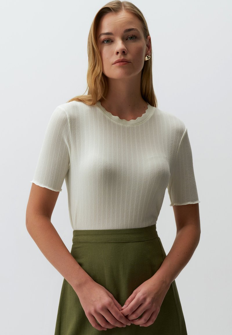 Biały prążkowany top z krótkim rękawem i falbaniastym dekoltem oraz lekko strzępionymi krawędziami, w połączeniu z zieloną spódnicą o wysokim stanie.