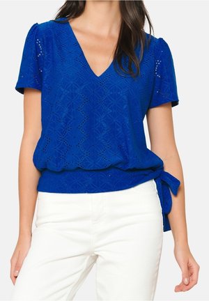 Niebieska bluzka z krótkim rękawem w dekoltem w serek, z kwiatowym wzorem ażurowym i wiązaniem z boku, noszona z białymi spodniami.