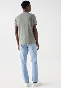 Šedé tričko s krátkým rukávem a centrálním švem, v kombinaci s světle modrými rovnými džíny a bílými teniskami, viděné ze zadní strany.