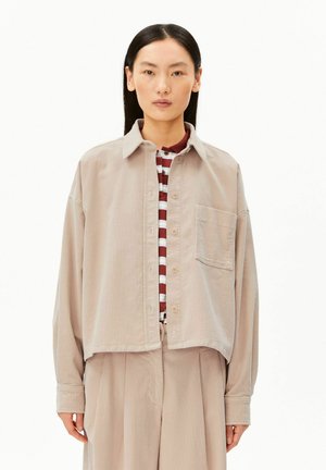 Vrouw met stijl zwart haar draagt een beige corduroy blouse met knopen en bijpassende broek over een bordeauxrood-wit gestreept topje.