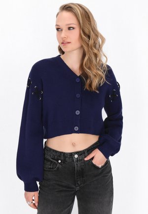 Kvinde med bølget blondt hår iført en marineblå cropped cardigan med knaplukning og sorte jeans med høj talje, stående med den ene hånd i lommen.
