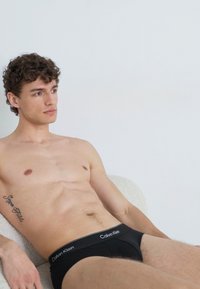 Schwarze Calvin Klein Unterhose mit elastischem Bund. Glattes Material und figurschmeichelndes Design. Model sitzt vor einem hellen Hintergrund.