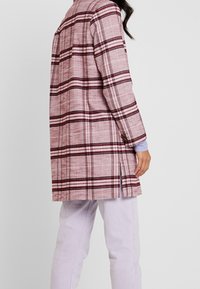 Kariert langer Mantel in Pink mit dunkelroten Streifen, strukturierter Stoff, seitliche Schlitze und traditioneller Kragen, kombiniert mit helllila Hosen.