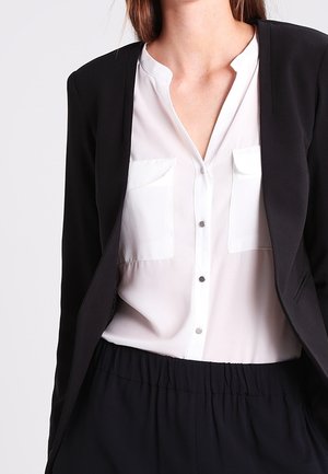 Kvinde iført en hvid skjorte med knapper og brystlommer under en sort blazer, kombineret med sorte bukser med høj talje.